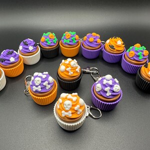 K&ouml;nnte beinhalten: Eine Sammlung von Halloween-Cupcake-Schl&uuml;sselanh&auml;ngern. Jeder Cupcake hat eine unterschiedlich farbige Glasur, darunter Lila, Orange und Wei&szlig;, und ist mit Totenk&ouml;pfen, Knochen und K&uuml;rbissen verziert. Die Cupcakes sitzen in verschiedenfarbigen Papierf&ouml;rmchen.