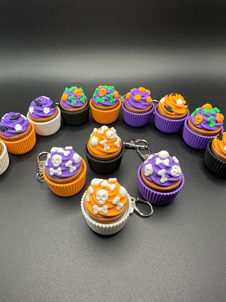 K&ouml;nnte beinhalten: Eine Sammlung von Halloween-Cupcake-Schl&uuml;sselanh&auml;ngern. Jeder Cupcake hat eine unterschiedlich farbige Glasur, darunter Lila, Orange und Wei&szlig;, und ist mit Totenk&ouml;pfen, Knochen und K&uuml;rbissen verziert. Die Cupcakes sitzen in verschiedenfarbigen Papierf&ouml;rmchen.