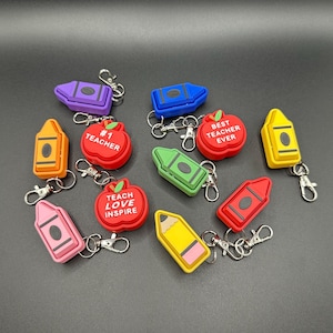 May include: Assortment of colorful teacher appreciation keychain charms. The charms feature a red apple with the text "#1 Teacher", a red apple with the text "Best Teacher Ever", a red apple with the text "Teach Love Inspire", a yellow crayon, a green crayon, a blue crayon, a purple crayon, a pink crayon, a red crayon, a yellow crayon with a pink eraser, and a yellow crayon with a black eraser. Each charm has a silver key ring.