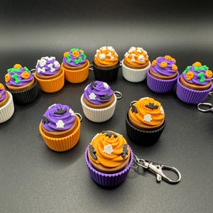 K&ouml;nnte beinhalten: Eine Sammlung von Halloween-Cupcake-Schl&uuml;sselanh&auml;ngern. Jeder Cupcake hat eine braune Basis, farbenfrohes Frosting und dekorative Elemente wie Flederm&auml;use, Geister und K&uuml;rbisse. Die Cupcakes sind in verschiedenen Farben erh&auml;ltlich, darunter Orange, Lila und Wei&szlig;.