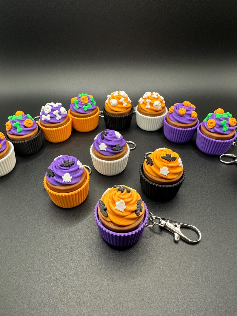 K&ouml;nnte beinhalten: Eine Sammlung von Halloween-Cupcake-Schl&uuml;sselanh&auml;ngern. Jeder Cupcake hat eine braune Basis, farbenfrohes Frosting und dekorative Elemente wie Flederm&auml;use, Geister und K&uuml;rbisse. Die Cupcakes sind in verschiedenen Farben erh&auml;ltlich, darunter Orange, Lila und Wei&szlig;.