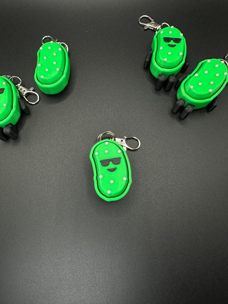 May include: A collection of green pickle-shaped keychains with white polka dots and black sunglasses. Each keychain has a silver clasp. The keychains are arranged on a dark gray surface.