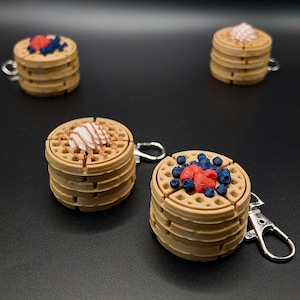 May include: Four waffle-shaped keychains with silver-colored clasps. Each keychain is a stack of waffles, with toppings of red and blue berries, and white cream. The waffles are light brown.