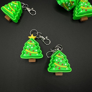 May include: Green Christmas tree keychains with yellow and red accents. Each keychain has a silver clasp and a brown base. The keychains are scattered on a black surface.