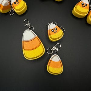 May include: Candy corn-shaped keychains in various sizes. The keychains feature the classic tri-color design of white, orange, and yellow, with a smiling face on the yellow base. Each keychain has a silver-colored clasp for attaching to keys or bags.