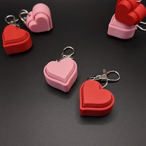 May include: Several heart-shaped keychains in shades of pink and red are scattered on a dark surface. Each keychain features a layered heart design and a silver-colored clasp. The keychains are a romantic accessory.