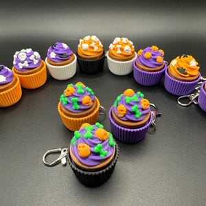 K&ouml;nnte beinhalten: Eine Sammlung von Halloween-Cupcake-Schl&uuml;sselanh&auml;ngern. Jeder Cupcake hat eine braune Basis, eine wei&szlig;e oder farbige Form und ist mit lila, orangefarbenem oder wei&szlig;em Zuckerguss bedeckt. Dekorationen umfassen Totenk&ouml;pfe, Knochen und K&uuml;rbisse.