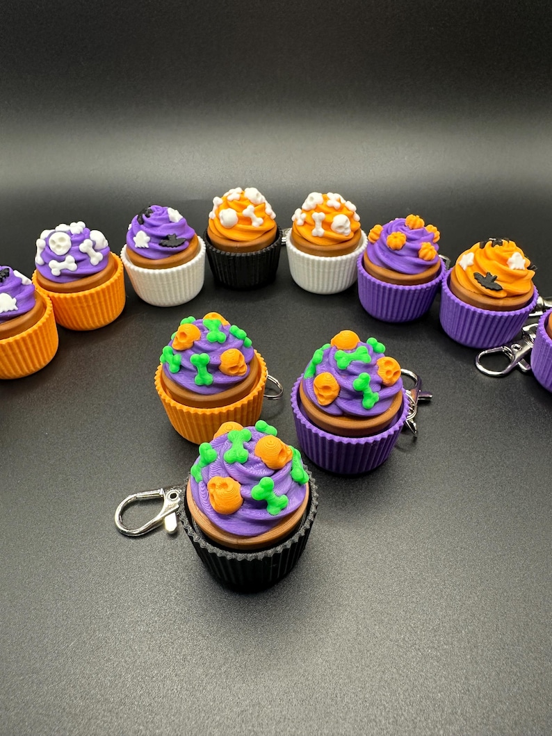 K&ouml;nnte beinhalten: Eine Sammlung von Halloween-Cupcake-Schl&uuml;sselanh&auml;ngern. Jeder Cupcake hat eine braune Basis, eine wei&szlig;e oder farbige Form und ist mit lila, orangefarbenem oder wei&szlig;em Zuckerguss bedeckt. Dekorationen umfassen Totenk&ouml;pfe, Knochen und K&uuml;rbisse.