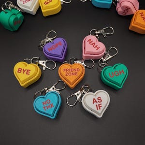 May include: A collection of heart-shaped keychains in various colors, each with a different phrase in white lettering. Phrases include "FRIEND ZONE", "AS IF", "NOPE", "NAH", "BYE", "NO THX", and "UGH". The keychains have silver-colored clasps.