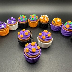K&ouml;nnte beinhalten: Eine Sammlung von Halloween-Cupcakes mit verschiedenen Dekorationen. Die Cupcakes haben lila, orangefarbenes und wei&szlig;es Frosting mit Designs wie K&uuml;rbissen, Flederm&auml;usen und Totenk&ouml;pfen. Die Cupcakes befinden sich in schwarzen, orangefarbenen und wei&szlig;en Cupcake-Formen.