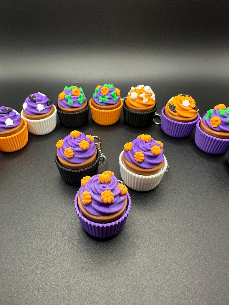 K&ouml;nnte beinhalten: Eine Sammlung von Halloween-Cupcakes mit verschiedenen Dekorationen. Die Cupcakes haben lila, orangefarbenes und wei&szlig;es Frosting mit Designs wie K&uuml;rbissen, Flederm&auml;usen und Totenk&ouml;pfen. Die Cupcakes befinden sich in schwarzen, orangefarbenen und wei&szlig;en Cupcake-Formen.
