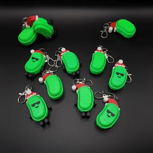 May include: A collection of green pickle-shaped keychains with black feet and sunglasses. Each keychain has a red and white Santa hat. The keychains are on a black surface.