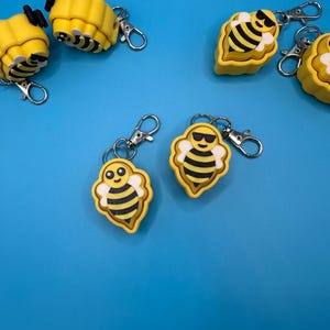 May include: Yellow bee-shaped keychains with black stripes and silver key rings. Some bees have sunglasses. The keychains are on a blue background. The keychains are made of a rubbery material.