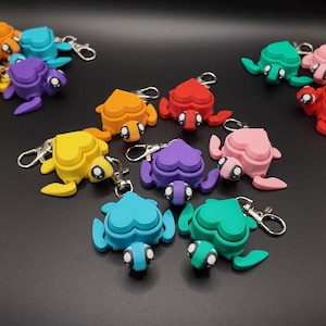 May include: A collection of colorful turtle-shaped keychains in various shades including blue, purple, yellow, orange, red, and green. Each keychain features a heart-shaped shell and a small, round head with black and white eyes. The keychains are made of a smooth material and have silver key rings.