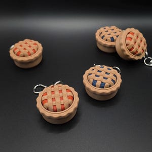 May include: Several miniature pie-shaped keychains. Each keychain features a tan crust with a lattice top and a filling of either red or blue. The keychains are on a black surface.