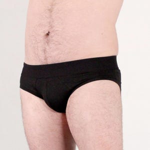 May include: Solid black briefs featuring a wide, elasticized waistband and a contoured front pouch. The text on the image reads: Xan, 5'7, 210lb, XL. These briefs provide full coverage and are designed with a simple, functional construction for daily wear.