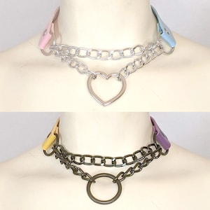 Op de afbeelding: Drie chokers met verschillende ontwerpen. De bovenste choker heeft een roze band, een zilveren ketting en een hartvormige hanger. De middelste choker heeft een gele band, een donkere ketting en een ringhanger. De onderste choker heeft een groene band, een zilveren ketting en een ringhanger.