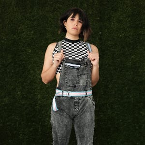 May include: A person wearing a black and white checkered crop top with a denim overall. The overall has a light blue and pink striped belt.
