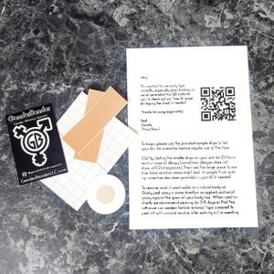 Puede incluir: Un logotipo en blanco y negro para Gender Bender Apparel con el texto "Gender Bender" y "Gender Binding Apparel" y un símbolo de una figura de género neutro. Una hoja de papel blanca con texto negro y un código QR. El texto incluye instrucciones para usar el producto y una nota para probar el producto en la piel antes de usarlo. El papel está al lado de dos tiras adhesivas beige y un pequeño círculo blanco.