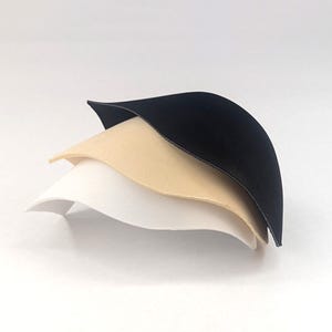 May include: A stack of three molded bra cups in black, beige, and white. Each cup features a smooth, rounded, semi-circular shape with a curved edge, designed to provide structure and support. The cups are layered on top of each other.