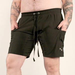 May include: Solid black shorts featuring an elastic waistband with a drawstring, two side pockets, and a side cargo-style pocket accented with two silver-toned metal grommets. The text on the image reads: Xan, 5'7, 210lb, XL.