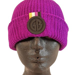 Puede incluir: Gorro de punto morado con un parche circular negro que dice "GB" en letras blancas. El parche tiene una pequeña franja amarilla, blanca y morada encima.