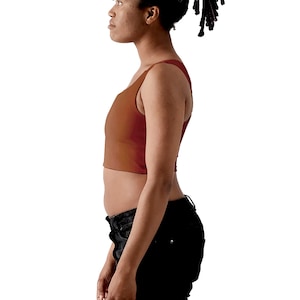 May include: A rust-colored, sleeveless crop top with a scoop neckline and a fitted silhouette. The garment is made of a smooth, solid-colored fabric that ends just above the waistline, providing a clean and simple look for casual wear.