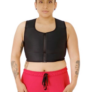 May include: Black compression top with a full-length zipper. The top is sleeveless and worn with red athletic pants with black side panels. The person in the image has short hair and tattoos on their arms.