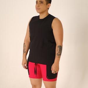 Puede incluir: Una persona lleva una camiseta negra sin mangas y pantalones cortos rojos con detalles negros. Los pantalones cortos tienen una cintura con cordón.