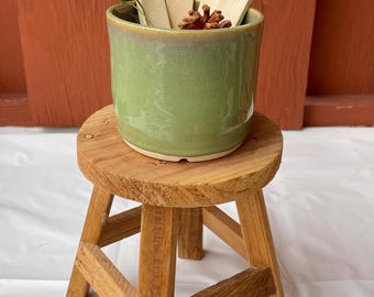 May include: A green ceramic pot with herb markers labeled Mint, Dill, Rosemary, and Sage, sits on a small wooden stool. The pot contains a small plant and dried herbs. The stool has a rustic, natural wood finish.