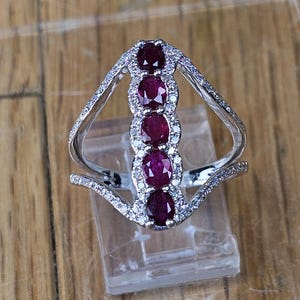 May include: A silver ring featuring five oval-cut, deep red rubies set vertically. The rubies are surrounded by small, clear accent stones. The ring has a unique, open design with curved bands.