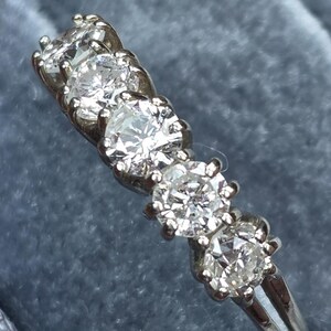 May include: A silver ring with five round, clear diamonds set in a row. The diamonds are held in place by small prongs. The ring is on a blue background.