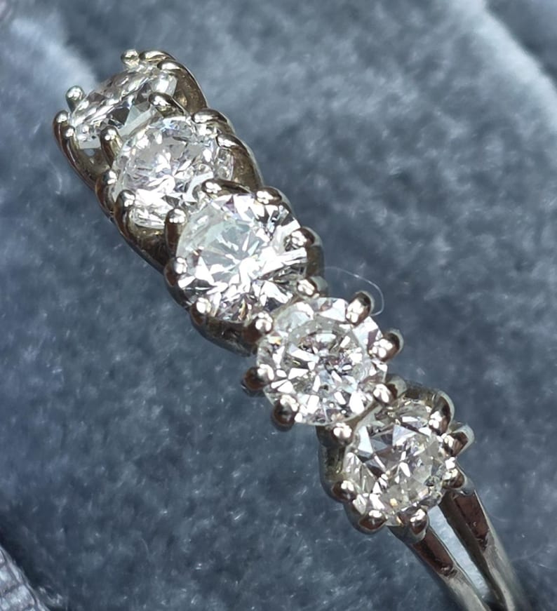 May include: A silver ring with five round, clear diamonds set in a row. The diamonds are held in place by small prongs. The ring is on a blue background.