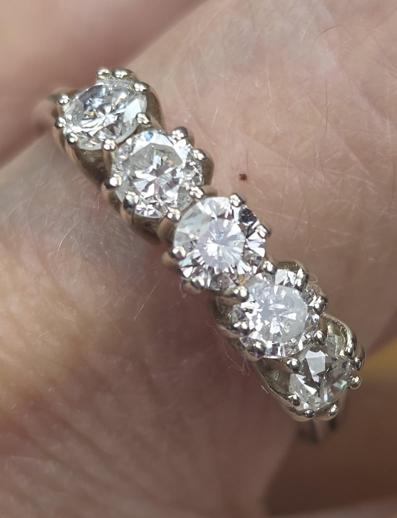 May include: A silver ring featuring five round, clear gemstones. The gemstones are set in individual, ornate settings, creating a sparkling, elegant design. The ring is likely made of silver or a similar metal.