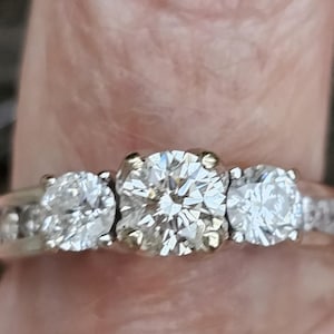 Puede incluir: Un anillo de compromiso de diamantes de tres piedras con una piedra central grande y dos piedras más pequeñas a cada lado. El anillo está engastado en oro blanco y tiene una banda de diamantes más pequeños.