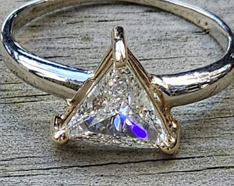 Könnte beinhalten: Ein silberner Ring mit einem dreieckigen, klaren Edelstein. Der Edelstein ist in einer goldfarbenen Lünette gefasst. Der Ring liegt auf einer verwitterten, grauen Holzoberfläche.
