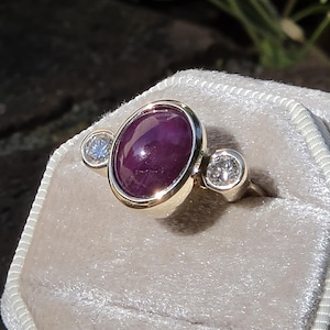 May include: A gold ring featuring a large, oval, deep red gemstone, flanked by two smaller, round, clear gemstones. The ring is set on a light gray velvet surface, with a blurred natural background.