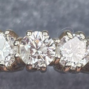 May include: A close-up of a five-stone diamond ring. The ring features five round-cut diamonds set in a white metal band. The diamonds sparkle brightly, reflecting light. The ring is set against a gray background.