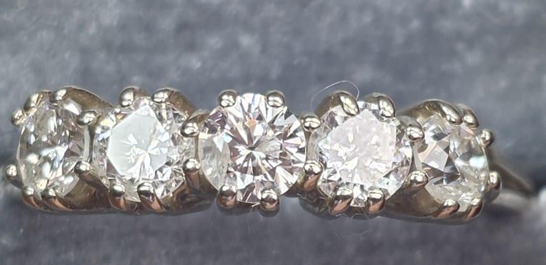 May include: A close-up of a five-stone diamond ring. The ring features five round-cut diamonds set in a white metal band. The diamonds sparkle brightly, reflecting light. The ring is set against a gray background.