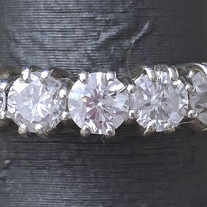May include: A close-up of a silver ring featuring five clear, round-cut diamonds. The diamonds are set closely together, creating a continuous sparkle. The ring is displayed against a dark background, highlighting the brilliance of the gemstones.