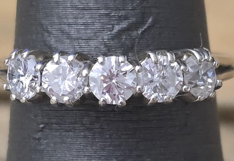 May include: A close-up of a silver ring featuring five clear, round-cut diamonds. The diamonds are set closely together, creating a continuous sparkle. The ring is displayed against a dark background, highlighting the brilliance of the gemstones.