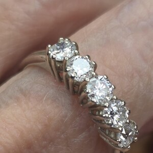 May include: A silver ring featuring five round, brilliant-cut diamonds. The diamonds are set in individual prongs, creating a sparkling display. The ring is shown on a finger, highlighting the jewelry's design and elegance.