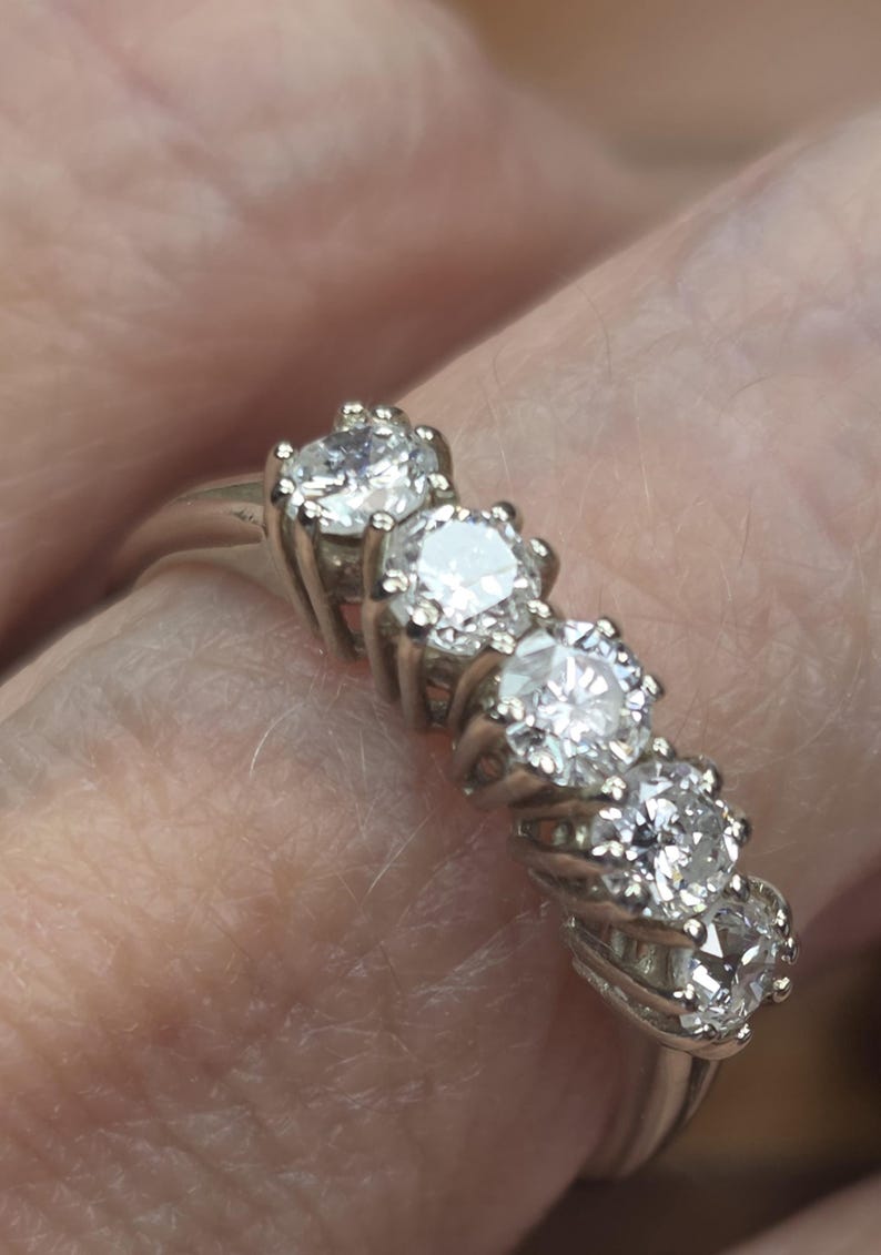 May include: A silver ring featuring five round, brilliant-cut diamonds. The diamonds are set in individual prongs, creating a sparkling display. The ring is shown on a finger, highlighting the jewelry's design and elegance.