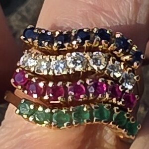 Puede incluir: Un anillo de oro con cuatro bandas onduladas apiladas, cada una engastada con una hilera de pequeñas piedras preciosas redondas. La banda superior contiene zafiros azul oscuro, la segunda diamantes, la tercera rubíes rojos y la inferior esmeraldas verdes.