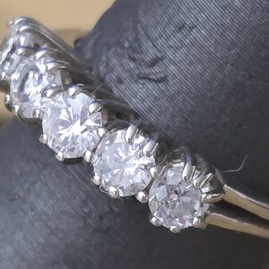 May include: Close-up of a silver ring with five clear, round-cut gemstones. The ring is set on a dark gray surface. The gemstones are evenly spaced and sparkle brightly.