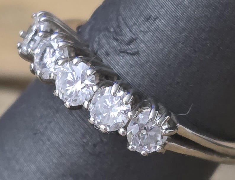 May include: Close-up of a silver ring with five clear, round-cut gemstones. The ring is set on a dark gray surface. The gemstones are evenly spaced and sparkle brightly.