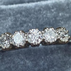 May include: A silver ring featuring five round, clear gemstones. The gemstones are set in individual prongs, and the ring is displayed against a soft, blue-gray background. The ring is a piece of jewelry.
