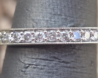 May include: A silver ring with a row of small, clear gemstones set in a channel. The ring has a delicate, beaded edge design. The ring is shown on a dark surface.