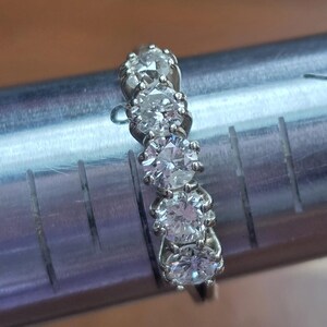 May include: A silver ring with five round, clear gemstones set in a row. The ring is on a metal ring sizer with measurements in inches. The gemstones are evenly spaced and appear to be diamonds.