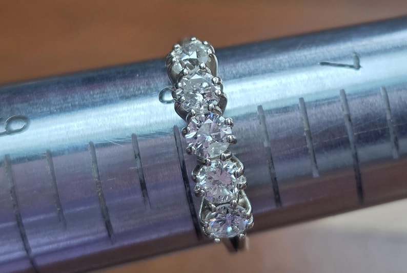 May include: A silver ring with five round, clear gemstones set in a row. The ring is on a metal ring sizer with measurements in inches. The gemstones are evenly spaced and appear to be diamonds.